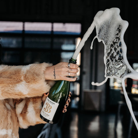 girl holding a bottle of three brothers wineries blanc de blanc shooting champagne from the cork