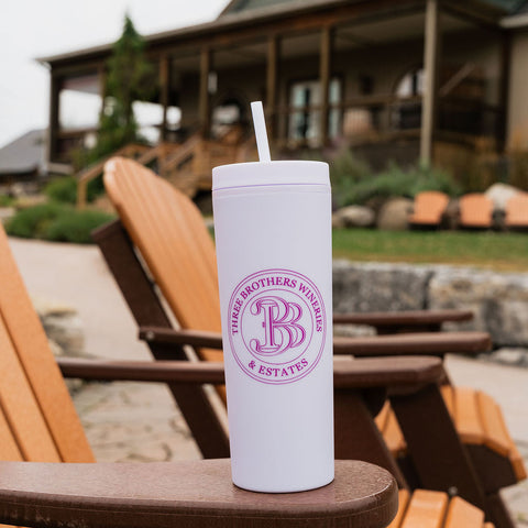 A white insulated tumbler with a straw sits on the arm of a wooden Adirondack chair in front of a house. The tumbler has a circular pink logo that reads 'Three BROTHERS VINTAGE & ESTATES' with a large monogram 3 B.