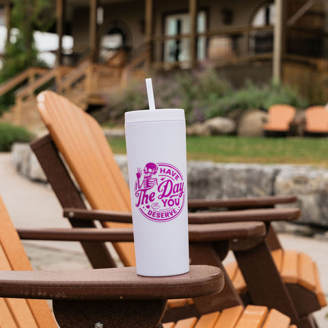 A white insulated tumbler with a straw sits on the arm of a wooden Adirondack chair in an outdoor patio setting, with a house and lawn blurred in the background. The tumbler features a purple circular graphic that reads 'Have THE Day YOU deserve'.