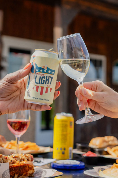 Two people toasting with a can of War Horse Light beer and a glass of white wine outdoors. Food in the background.