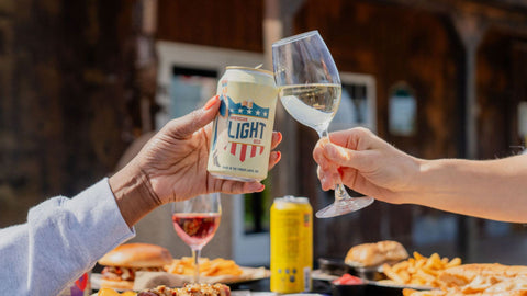 Two people toasting with a can of War Horse Light beer and a glass of white wine outdoors. Food in the background.